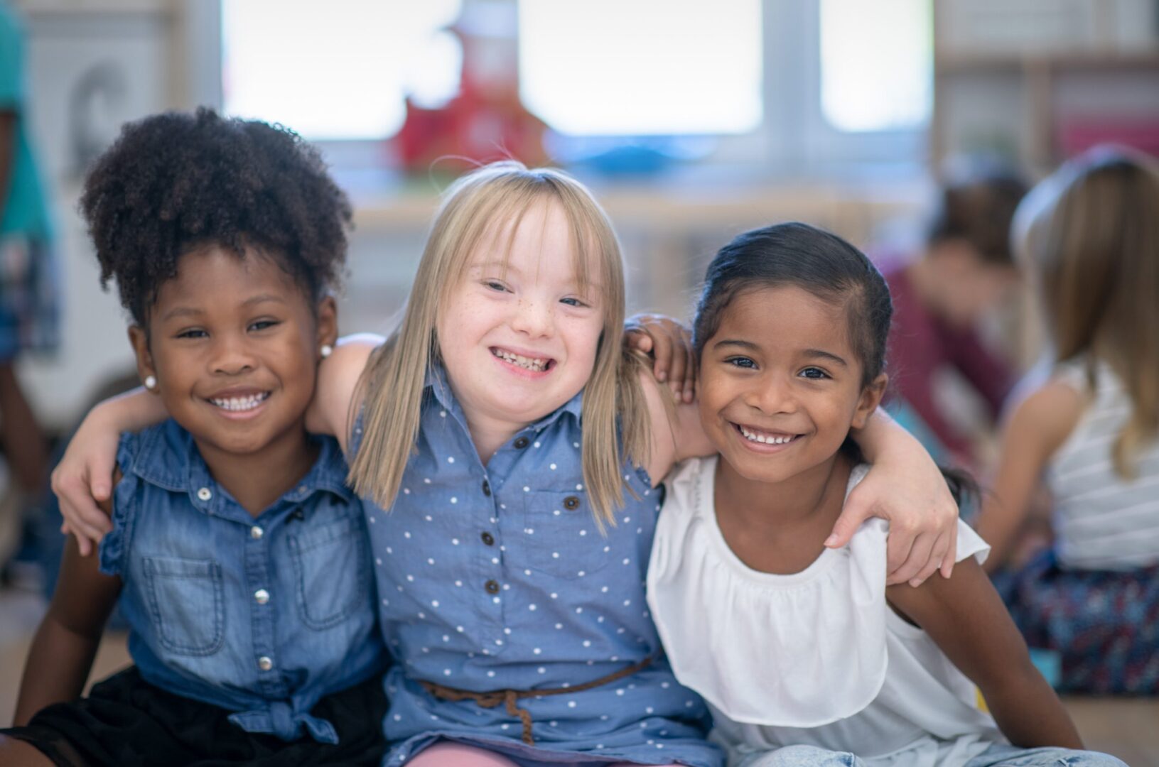 Three multi ethnic preschool girls in their classroom. One of the girls (in the middle) is of Caucasian ethnicity and has Down syndrome. The other girls are of African and mixed race ethnicity.